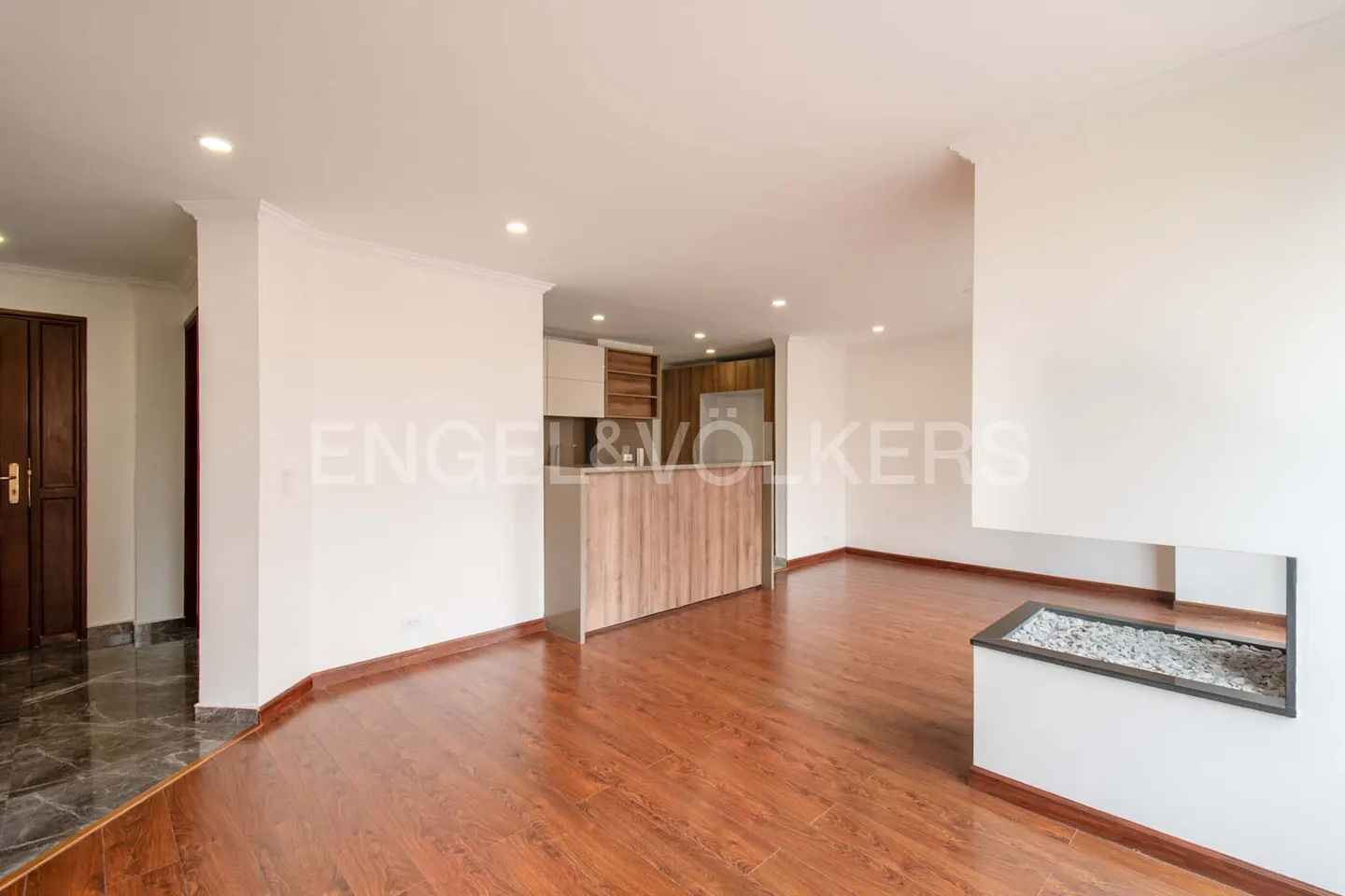 Open-concept living space with wood floors, white walls, and recessed lighting. A kitchen island separates the living area. A modern fireplace is on the right.