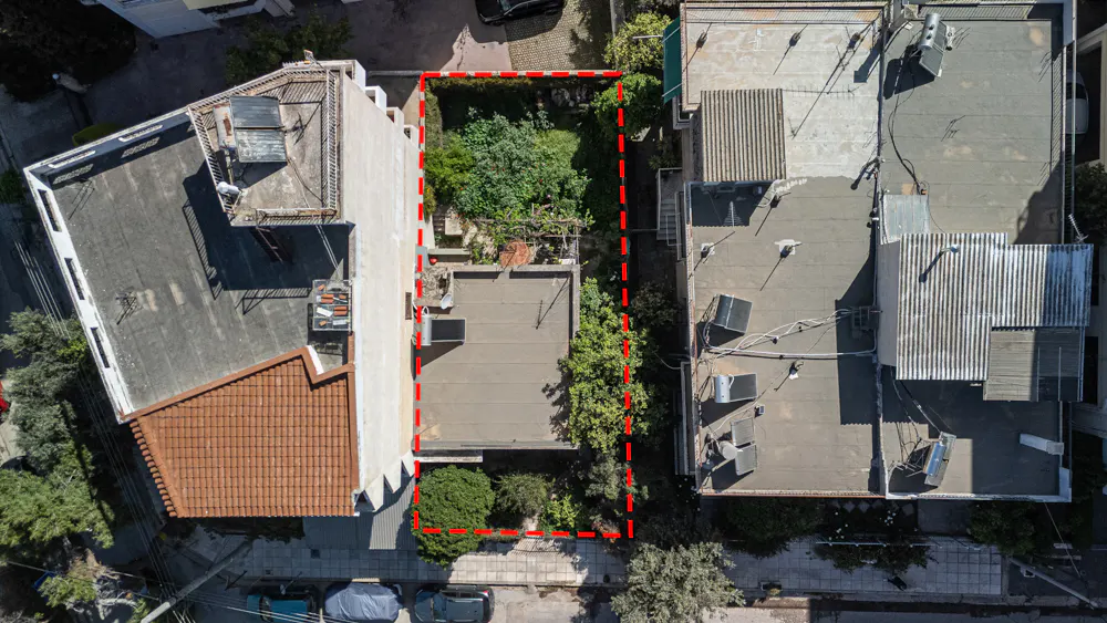 Aerial view of a property outlined in red, nestled between buildings with trees and greenery.