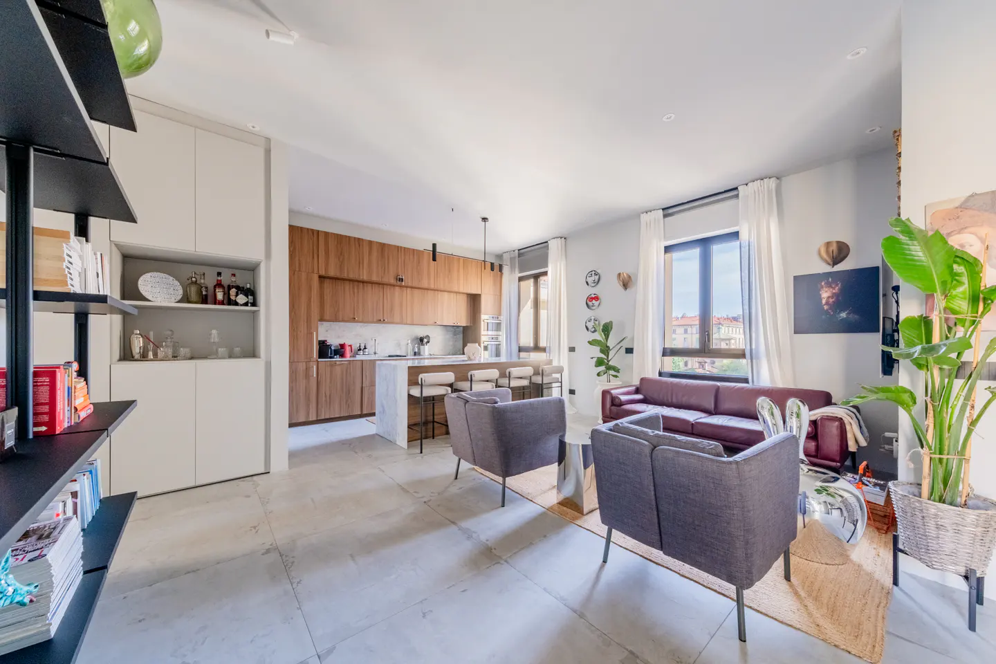 Modern open-plan living area with a burgundy sofa, two grey armchairs, a kitchen with wood cabinets, and a large plant.