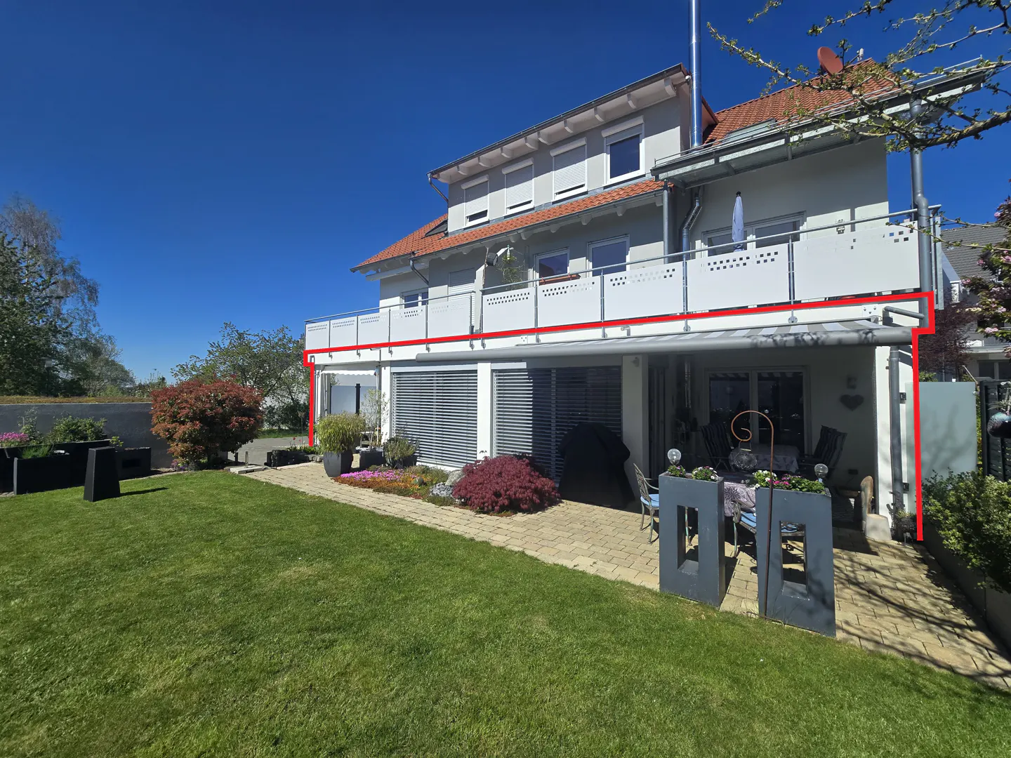 Two-story house with a white balcony, red roof, and a green lawn on a sunny day. A patio with furniture is visible.