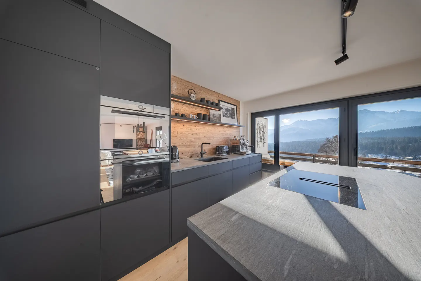 Modern kitchen with gray cabinets, stone countertops, and wood-paneled wall. Large windows offer a mountain view.