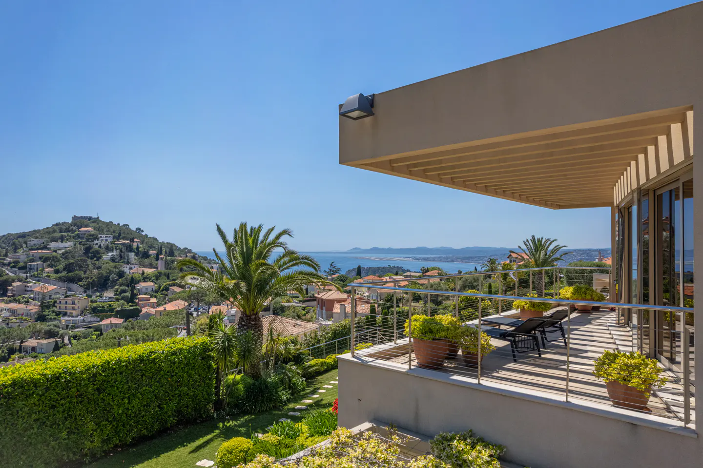Balcony view of a tan house overlooking a hillside town and ocean on a sunny day. Palm tree and potted plants on the balcony.