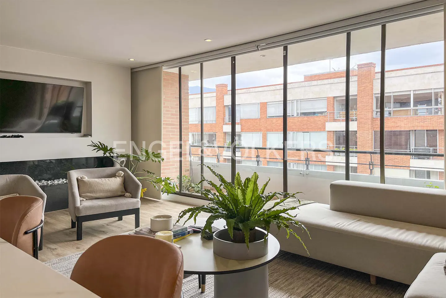 Bright living room with large windows showing a brick building. A white sofa, chairs, and a round table with a potted plant furnish the space.