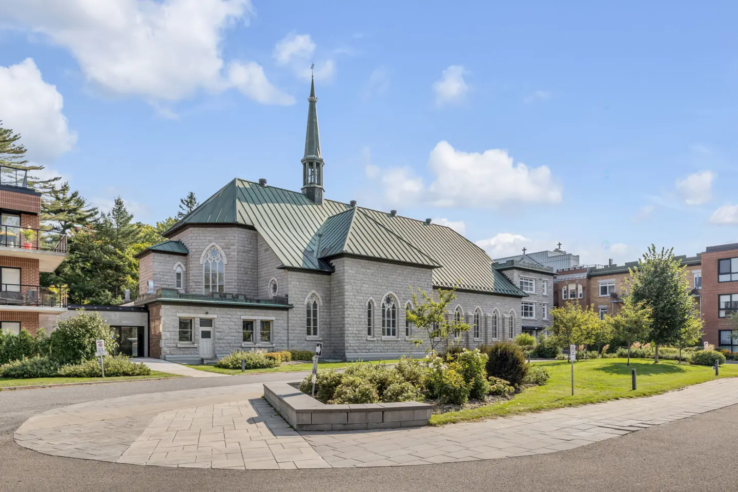 Exterior view of a gray stone church with a green roof and steeple, surrounded by green lawns and trees.