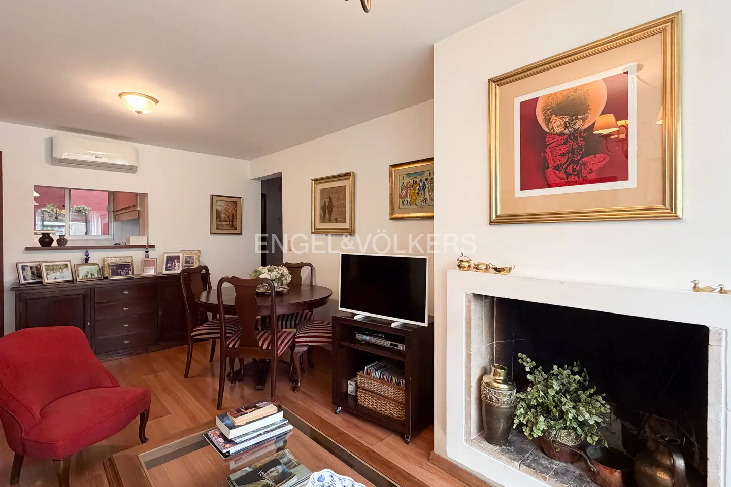 Living room with wood floors, a red chair, a dining table, and a fireplace with a gold-framed painting above it.