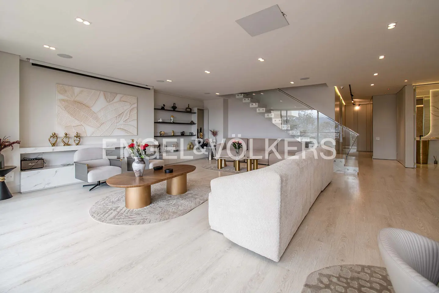 Bright, modern living room with white sofa, chairs, and wood floors. A glass staircase is visible in the background.