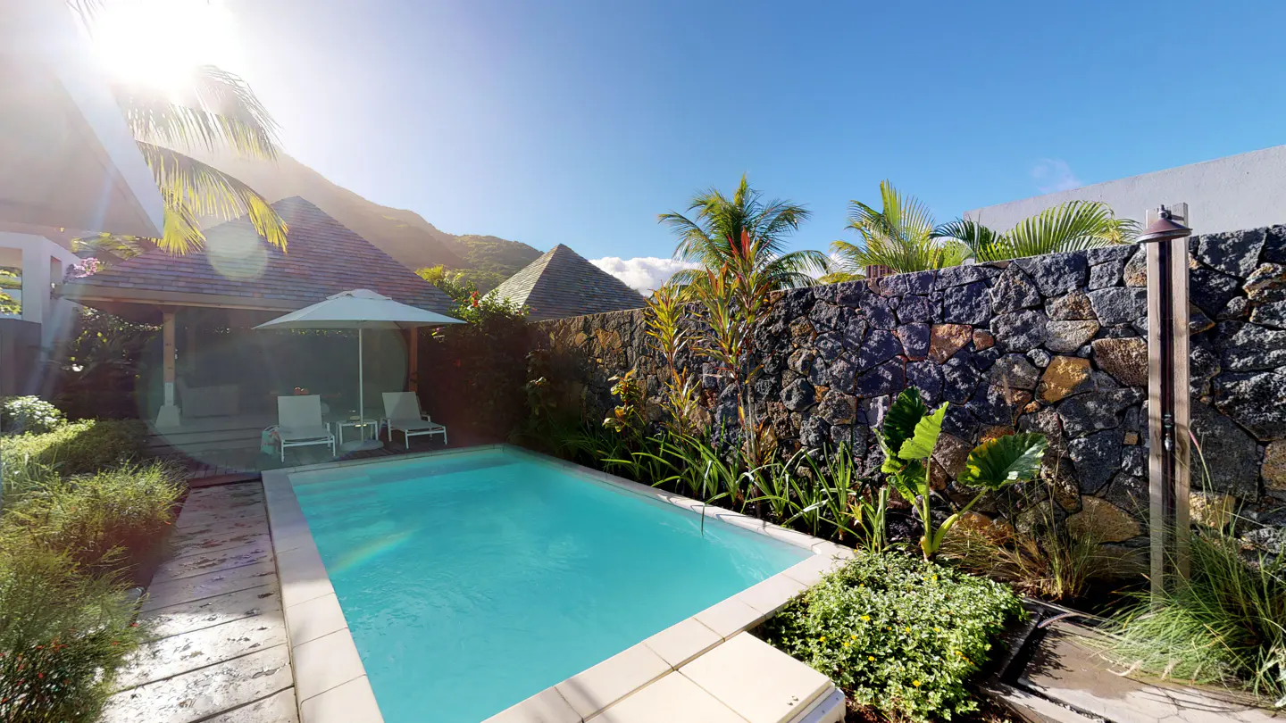 A turquoise pool is surrounded by lush greenery, a stone wall, and a cabana with lounge chairs under a white umbrella.