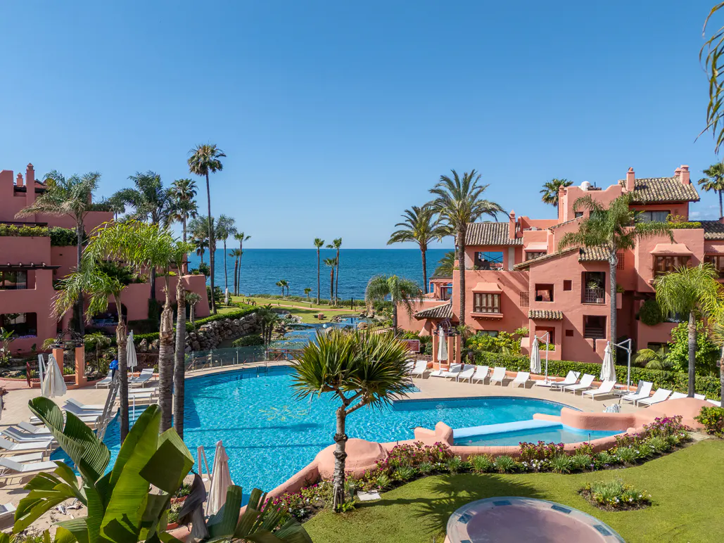 Resort view with a blue pool, palm trees, and pink buildings under a clear blue sky near the ocean.