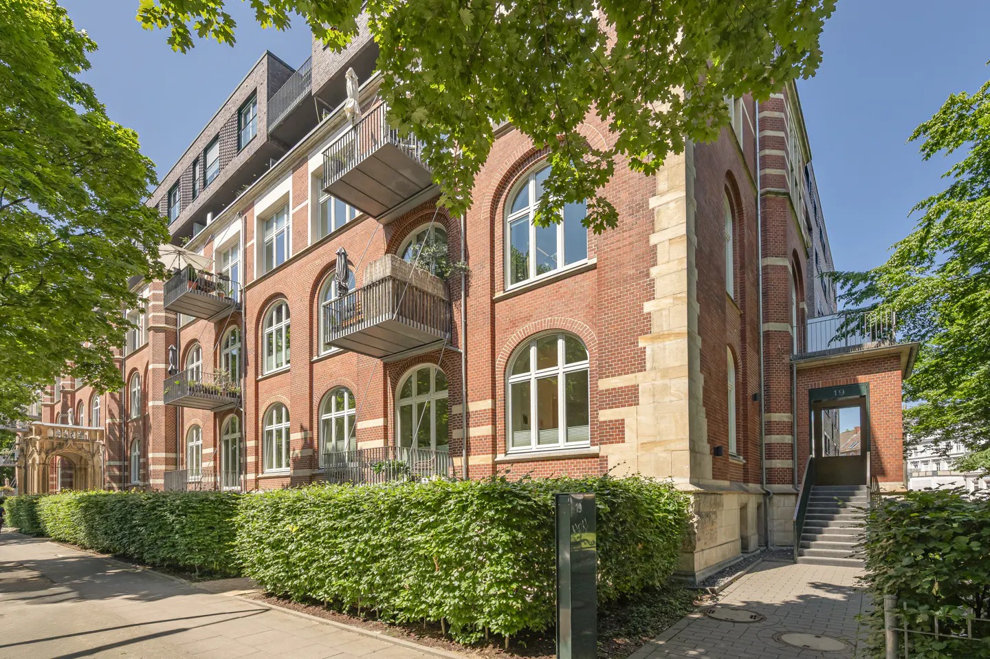 Exterior view of a red brick apartment building with arched windows and small balconies, surrounded by green trees and hedges.