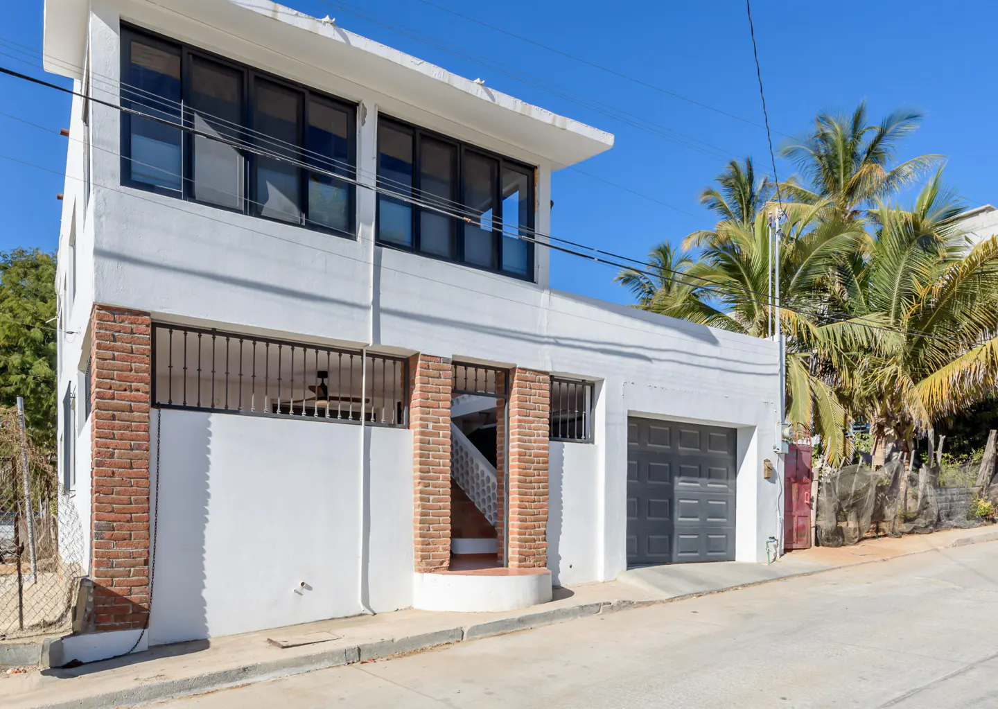 Exterior of a white, two-story house with a gray garage door and brick accents. Palm trees are visible in the background.