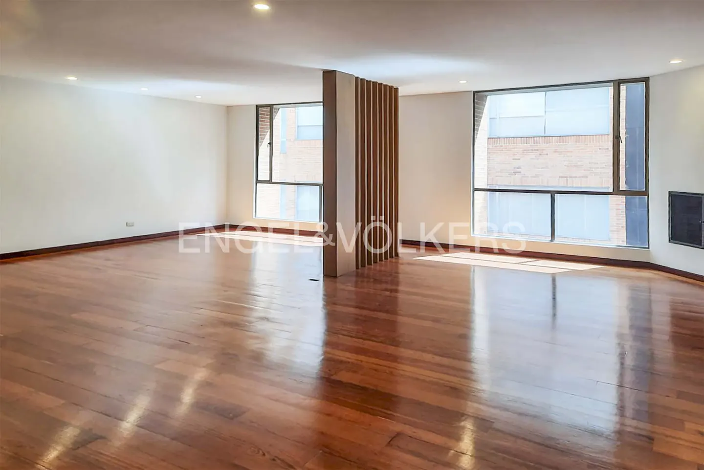 Empty room with hardwood floors, white walls, and large windows. A wooden slat divider stands in the center. Engel & Volkers logo is superimposed.