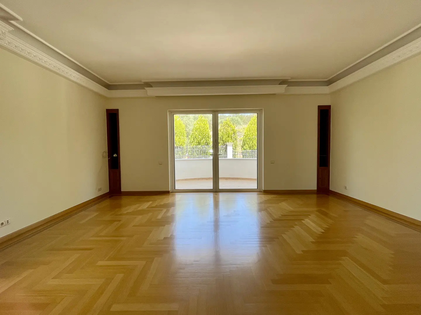Empty room with herringbone wood floor, cream walls, and white double doors to a balcony with green trees.