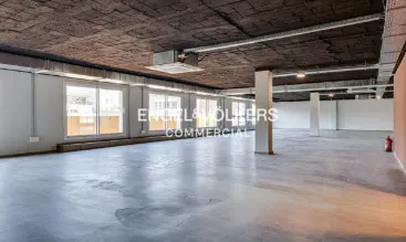 Empty commercial space with concrete floors and ceiling, white walls, and large windows. An Engel & Volkers logo is visible.