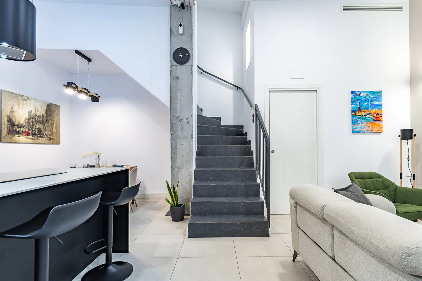 Modern open-plan living area with a black kitchen island, dark staircase, white walls, and a beige sofa with green chair.