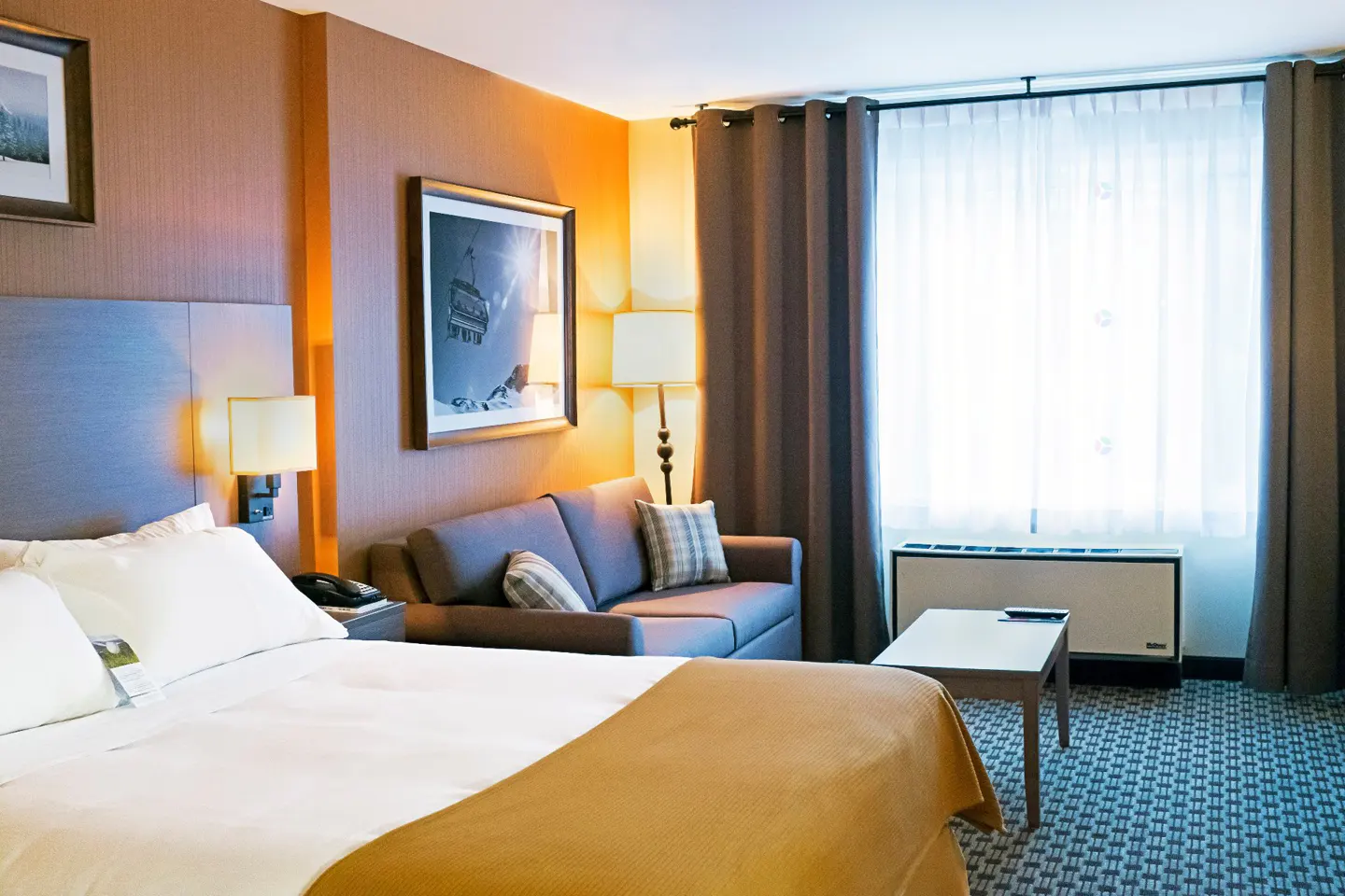 Hotel room with a king bed, brown sofa, and a window with sheer white curtains. A framed picture of a ski lift hangs on the wall.