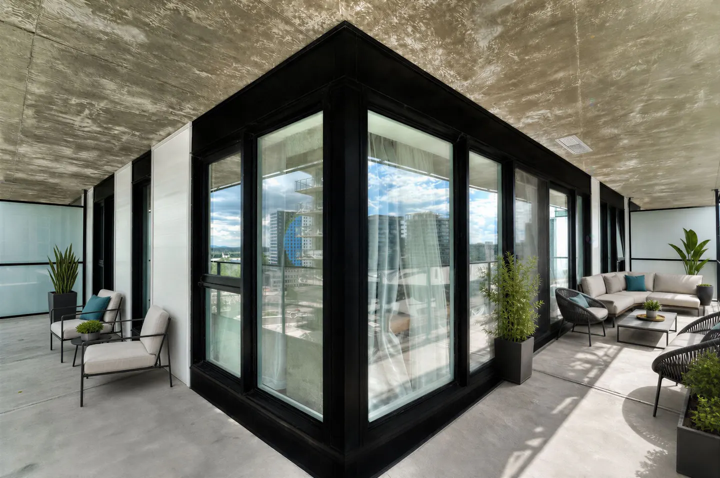 Wide angle view of a modern balcony with black framed windows, concrete floor and ceiling, and outdoor furniture. City view in background.