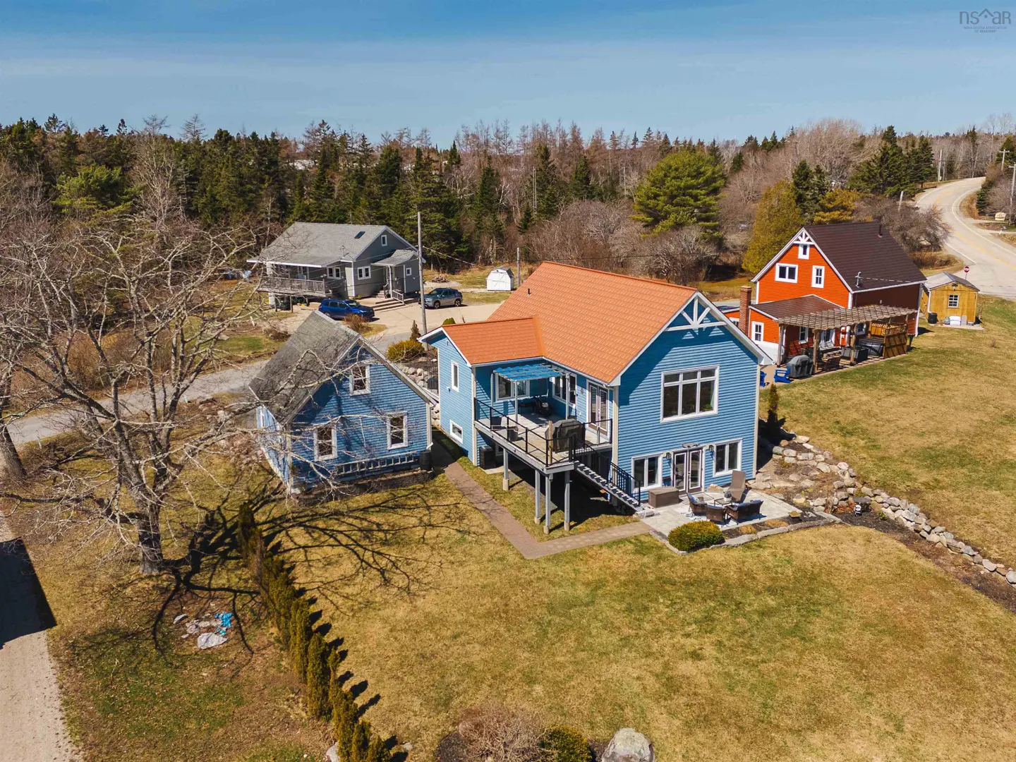 Aerial view of a blue house with an orange roof, a deck, and a patio with furniture. Other houses and trees are in the background.
