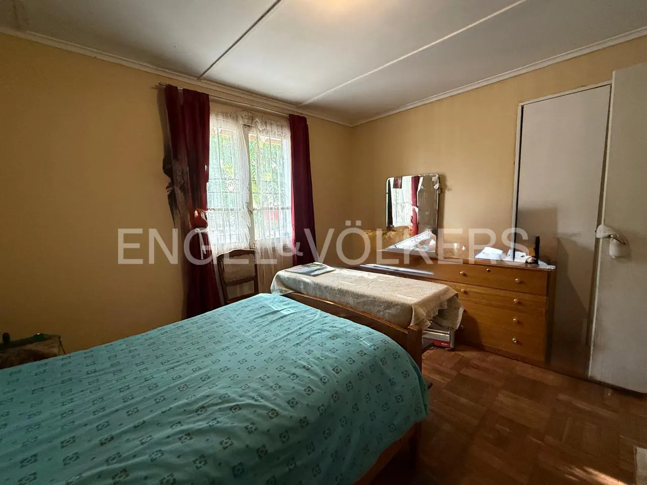 Bedroom with two beds, a wooden dresser with a mirror, and a window with red curtains.