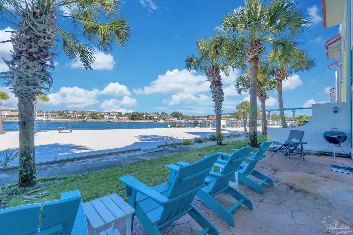 Beachfront patio with blue Adirondack chairs, palm trees, and a grill. White sand beach and blue water visible in the background.