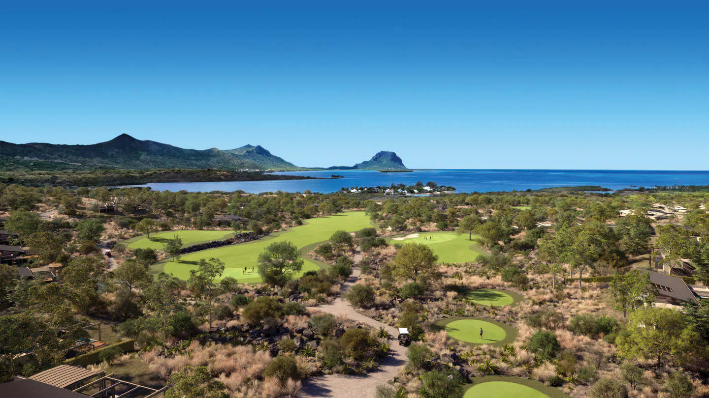 Aerial view of a lush green golf course with mountains and ocean in the background under a clear blue sky.