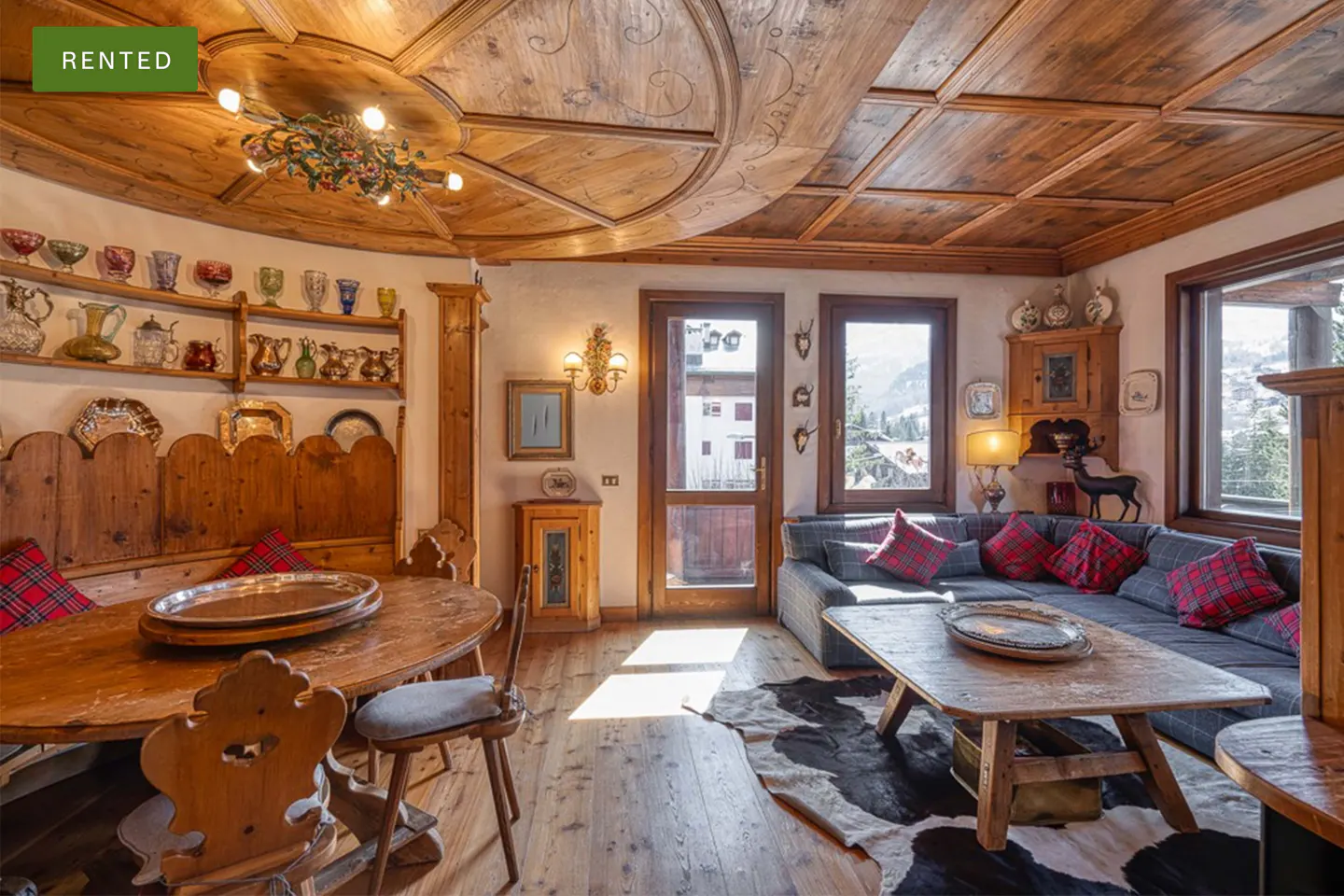 Rustic living room with wood-paneled ceiling, dining area, gray sofa with red plaid pillows, and a cowhide rug.