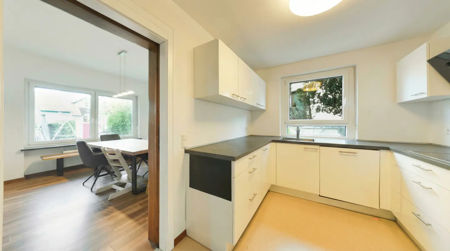 A bright kitchen with white cabinets and dark countertops, open to a dining room with a table and chairs.