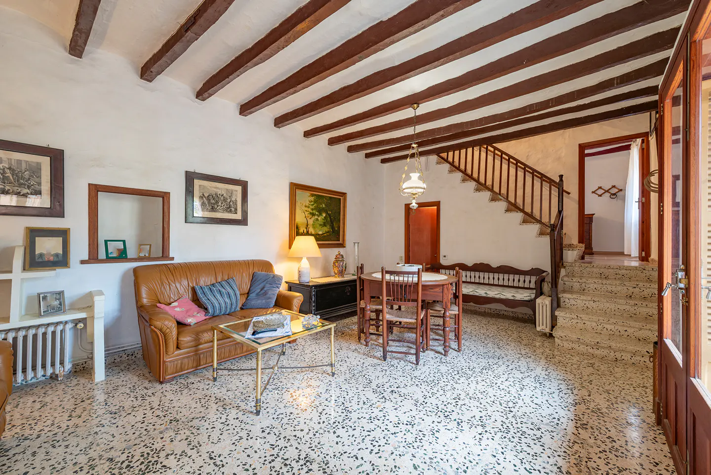 Living room with brown leather sofa, wood beams, and stairs. A table and chairs sit near the stairs. The floor is speckled.