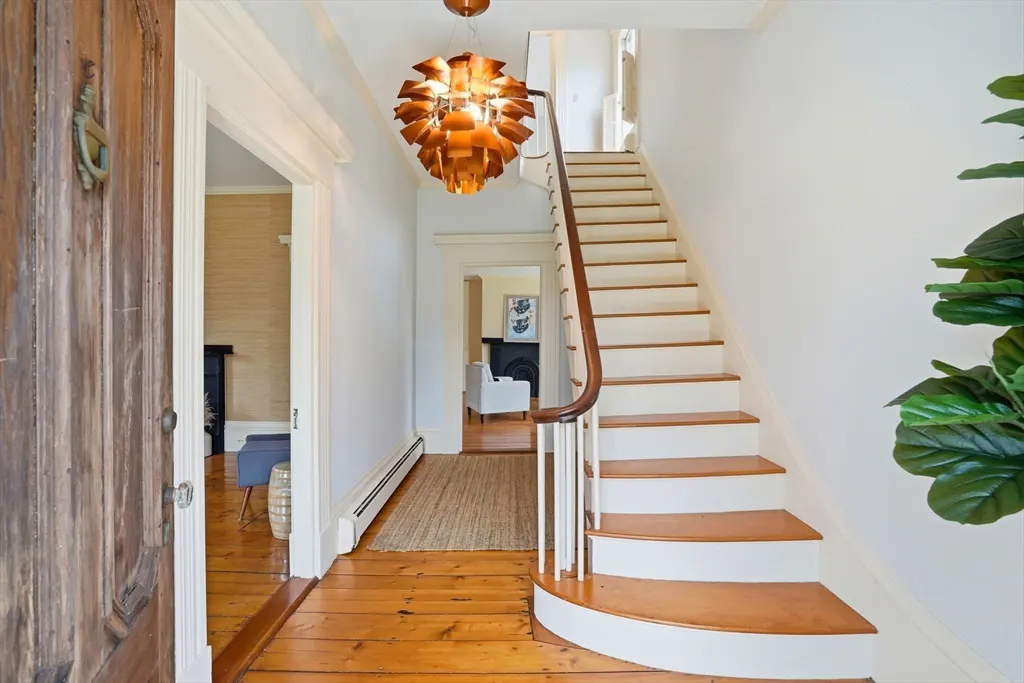 Bright foyer with wood floors, white walls, and staircase. A copper pendant light hangs above. An open door reveals a living room.