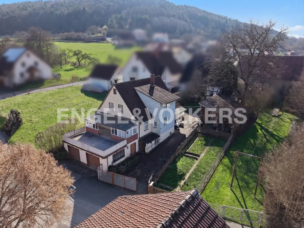 Aerial view of a two-story house with a brown roof, white walls, and a garage, surrounded by green grass and trees.
