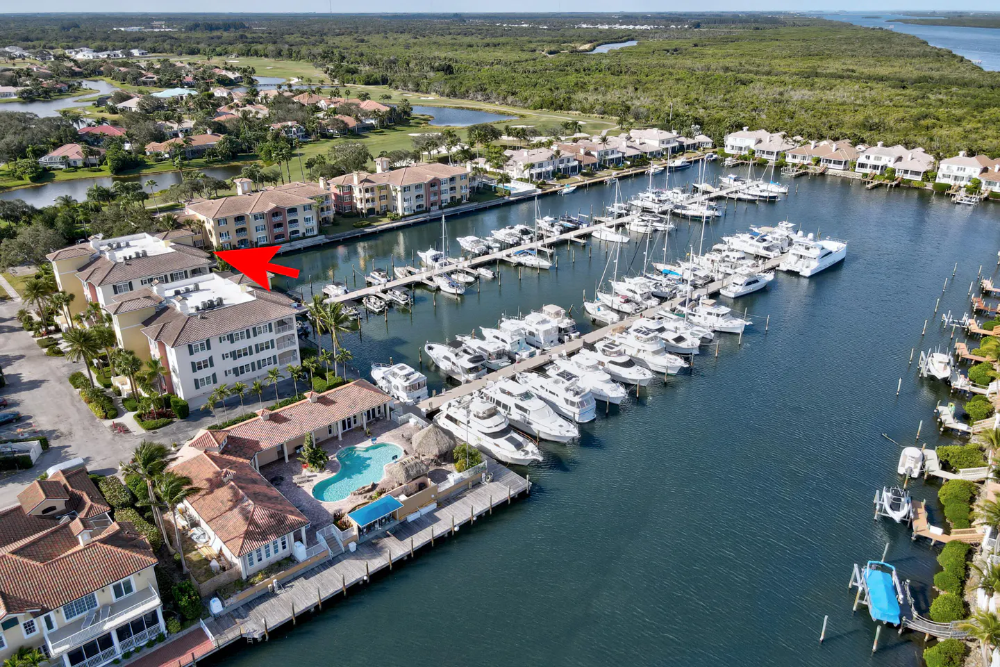 Aerial view of a waterfront condo complex with a marina, boats, and a pool. Red arrow points to a specific unit.