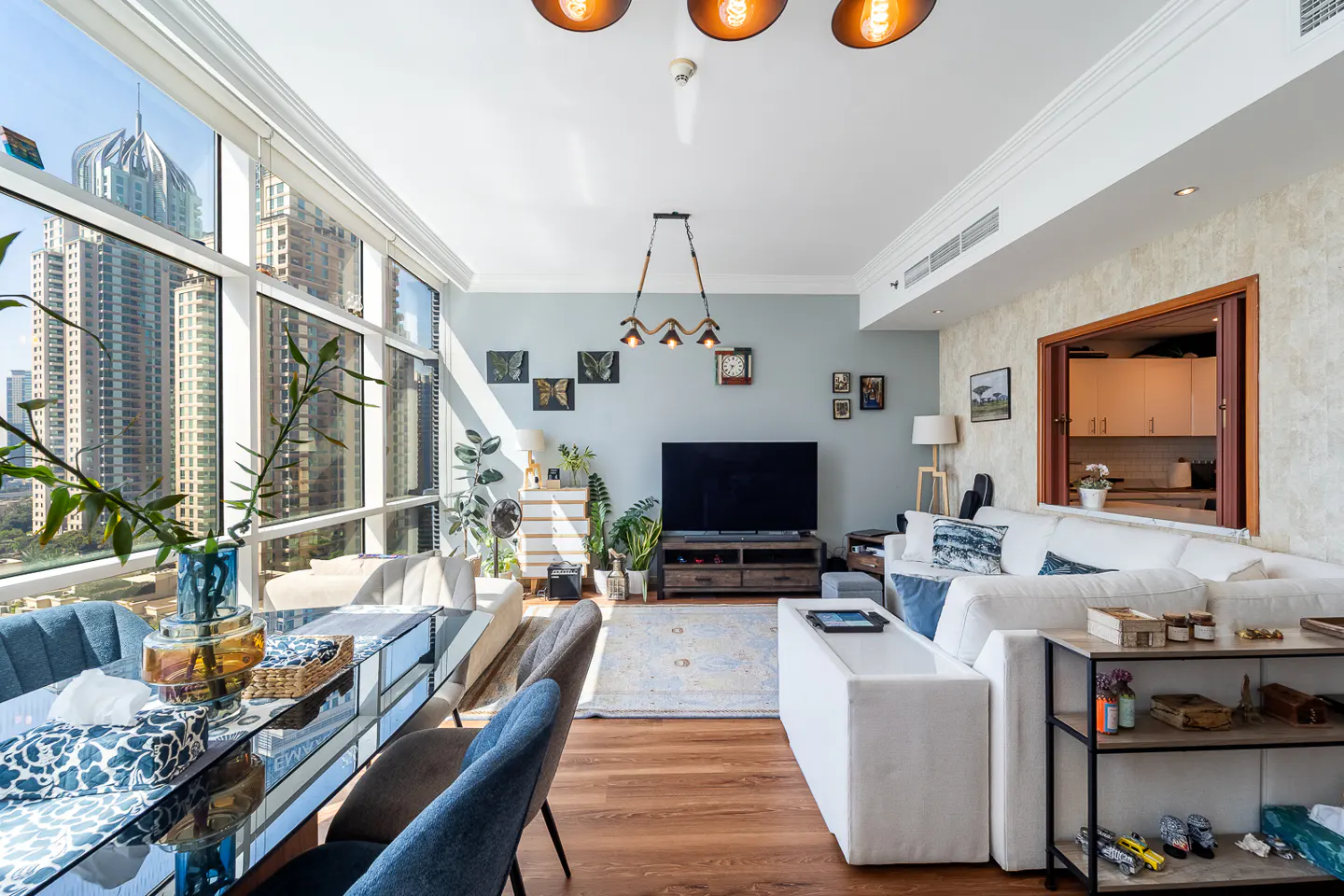 Bright living room with a glass dining table, blue chairs, and a white sofa. Large windows offer a city view. A TV and decor adorn the light blue walls.