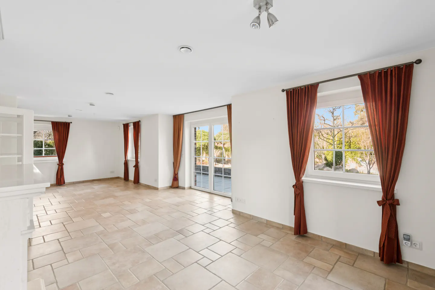 Bright, empty room with beige tile floor, white walls, and rust-colored curtains framing windows and glass doors.