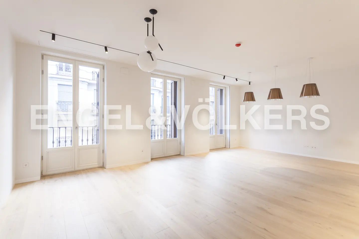 Bright, empty room with light wood floors, white walls, and three sets of white French doors with balconies. Modern lighting fixtures hang from the ceiling.