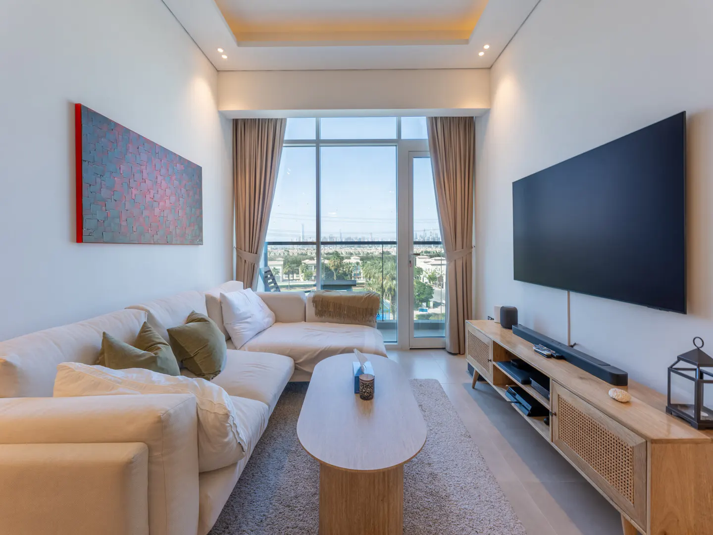 Living room with a white sectional sofa, oval coffee table, and a large TV. A modern art piece hangs on the wall. Large windows offer a city view.