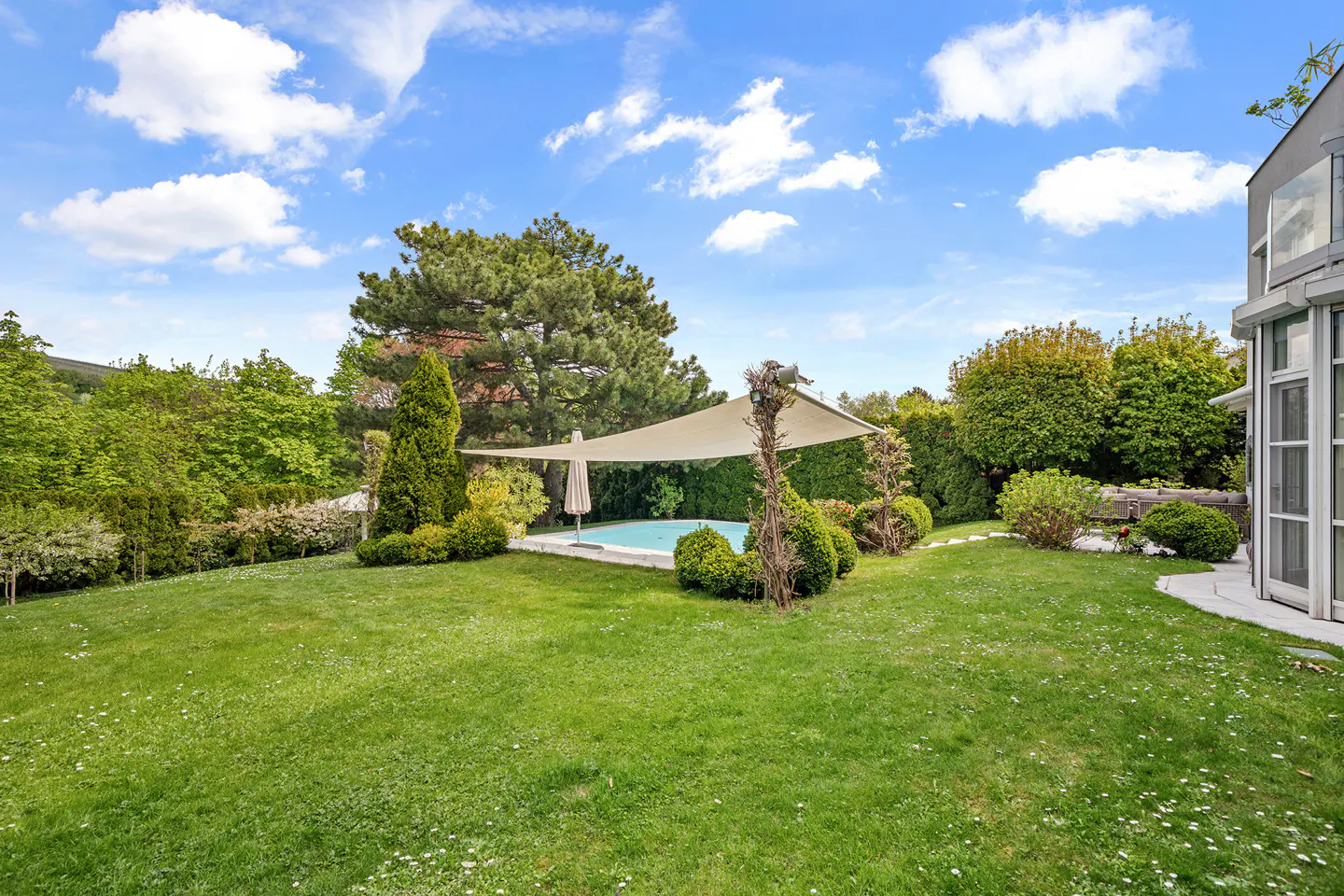 A backyard with a pool, sunshade, and green lawn under a blue sky with white clouds.