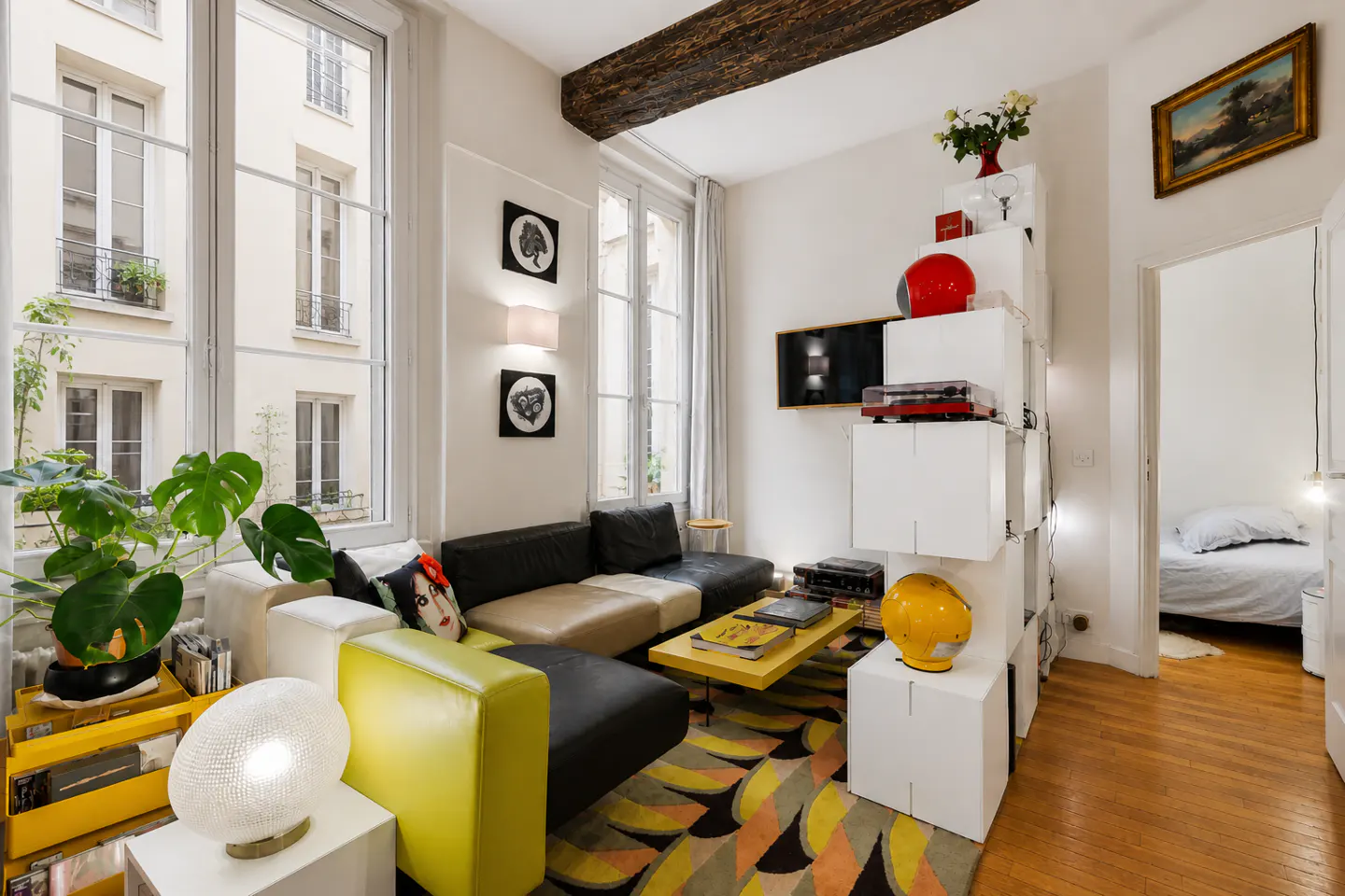 Bright living room with large windows, a black and tan sectional sofa, and a yellow coffee table on a patterned rug. A bedroom is visible through a doorway.