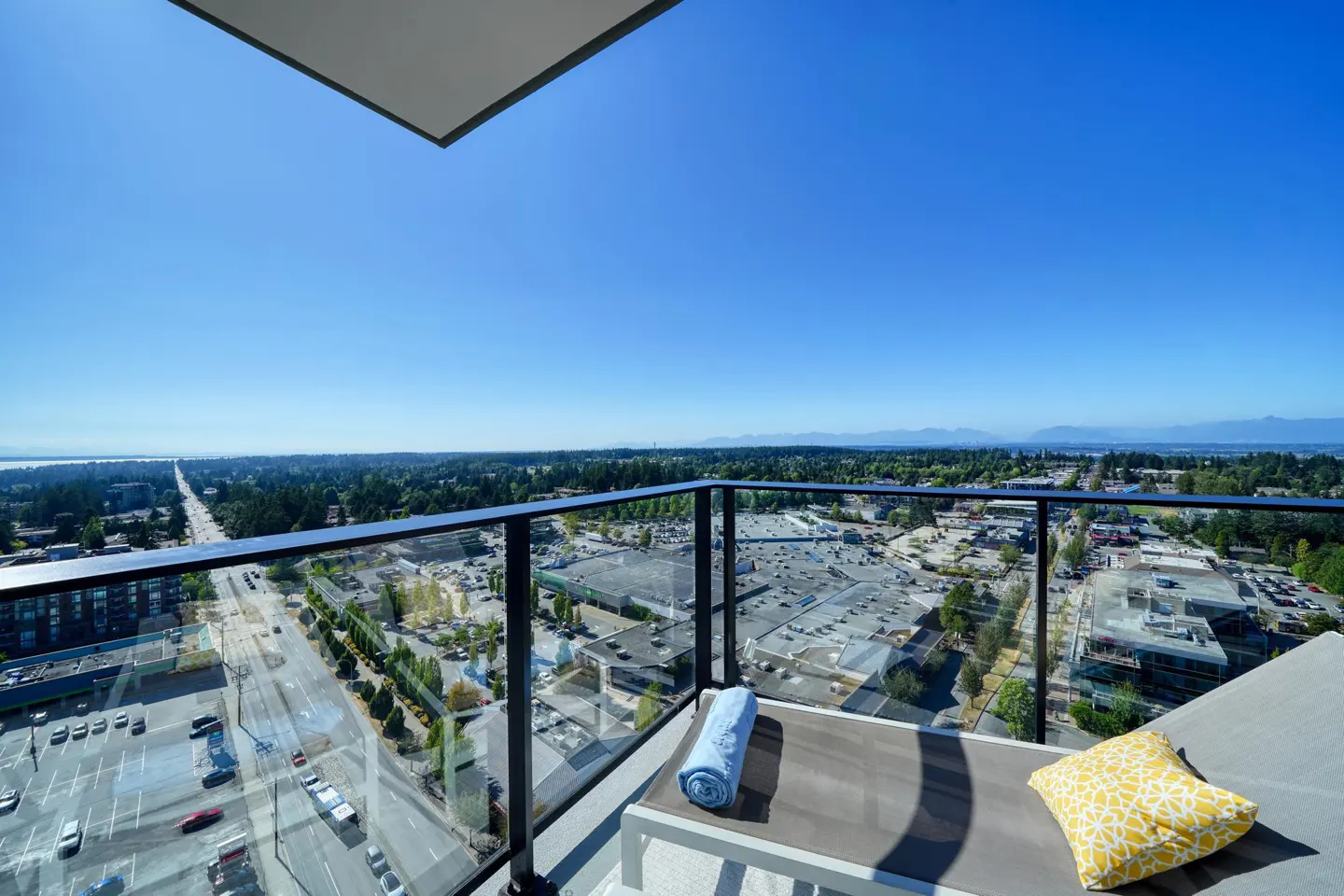 Balcony view with a lounge chair, blue towel, and yellow pillow. Cityscape with trees and mountains in the background under a clear blue sky.