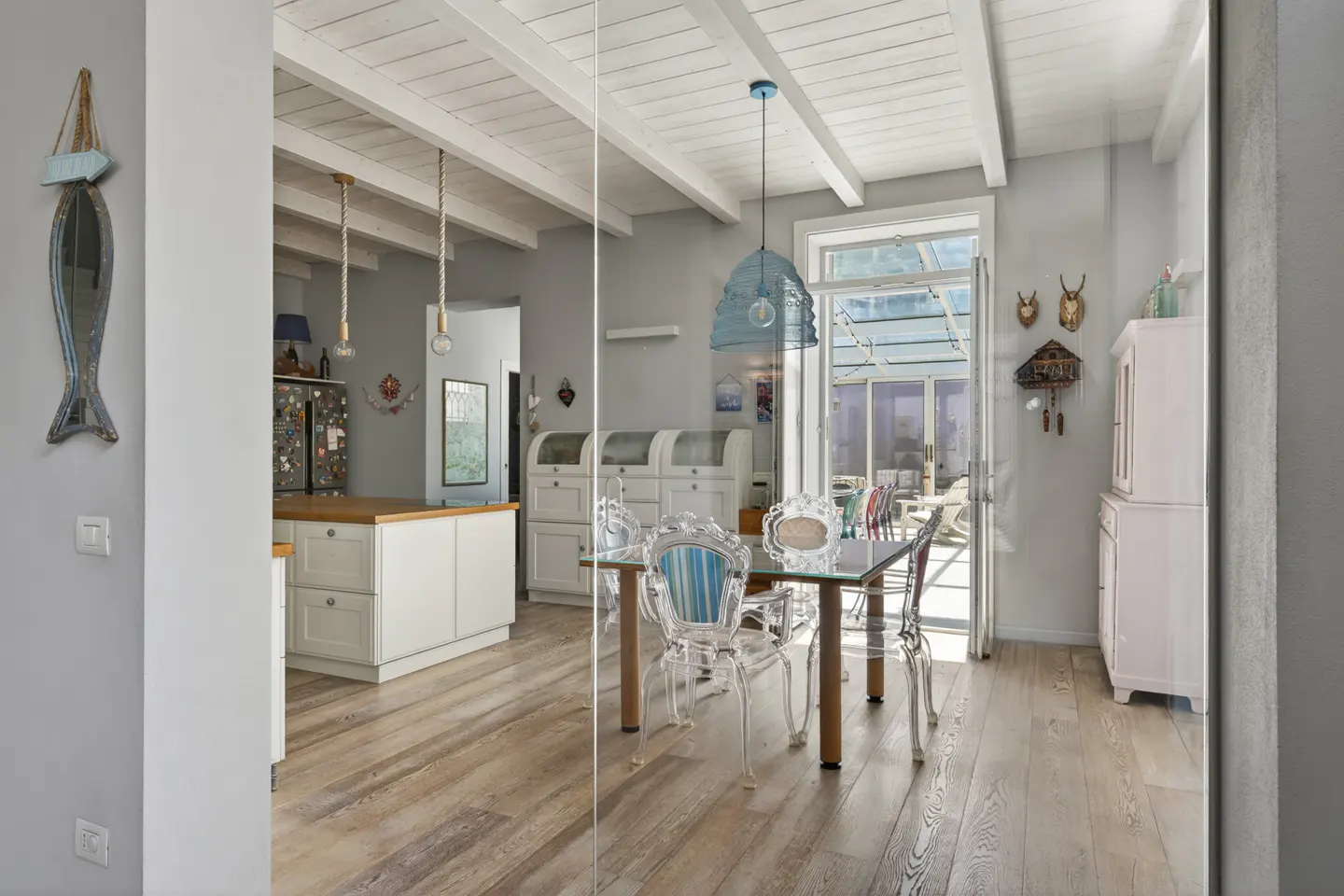Bright, airy dining room with wood floors, white cabinets, and a glass table with clear chairs. A blue fish mirror hangs on the wall.