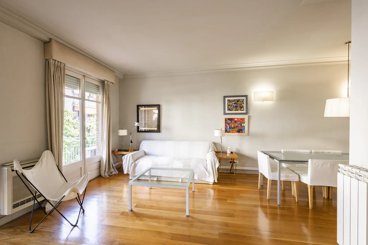Bright living room with hardwood floors, white sofa, butterfly chair, glass table, and dining set. Artwork adorns the light gray walls.