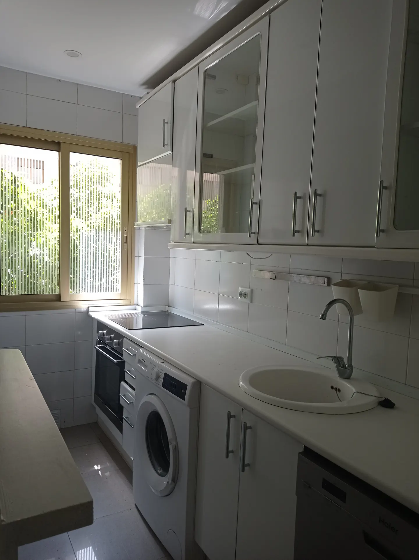 Bright kitchen with white cabinets, countertops, and appliances. A window with vertical blinds is on the left.