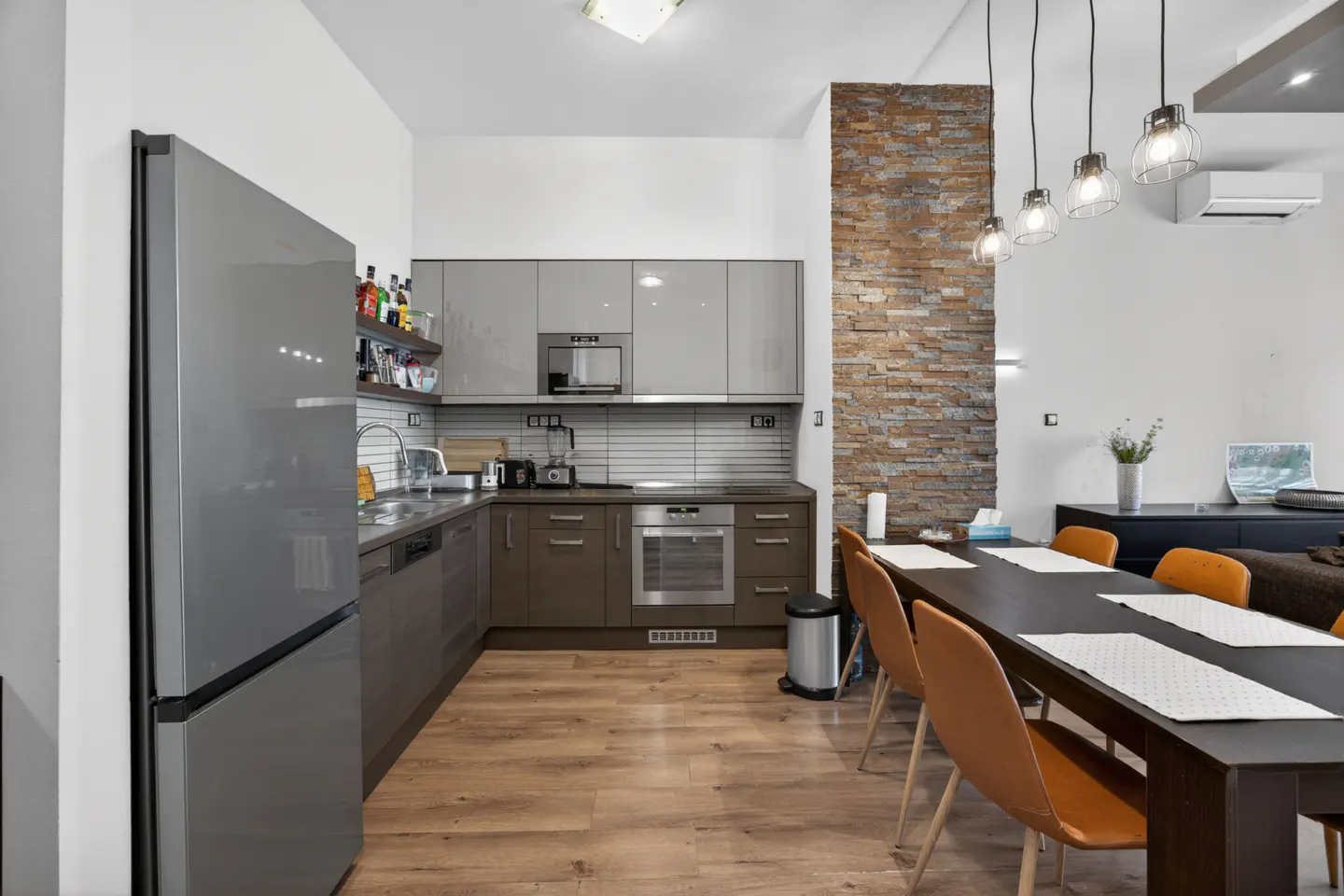 Modern kitchen with grey cabinets, stainless steel appliances, stone accent wall, and a dining table with orange chairs.