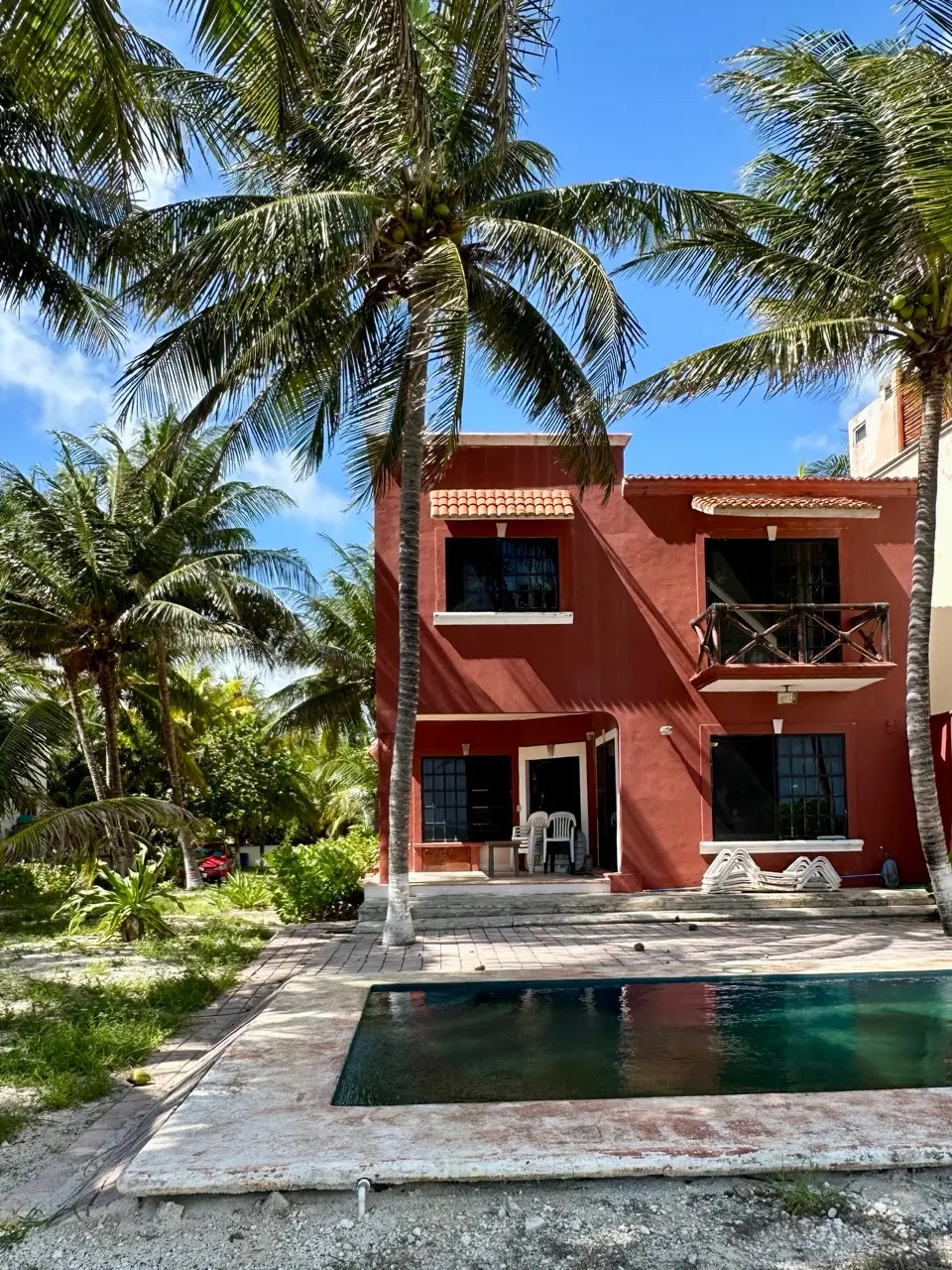 Two-story red house with a pool, surrounded by palm trees under a blue sky.