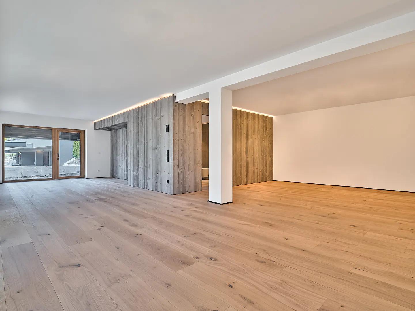 An empty room with light wood floors, white walls, and a concrete accent wall with recessed lighting. A glass door leads to the outside.