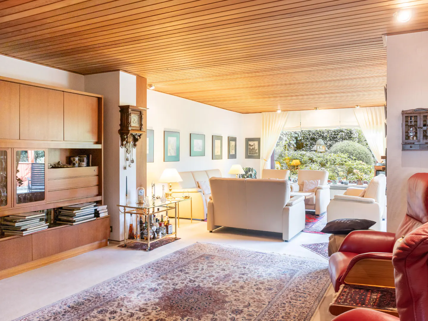 A living room with a wood ceiling, built-in shelves, and a view of a garden. Cream sofas and red leather chairs are visible.