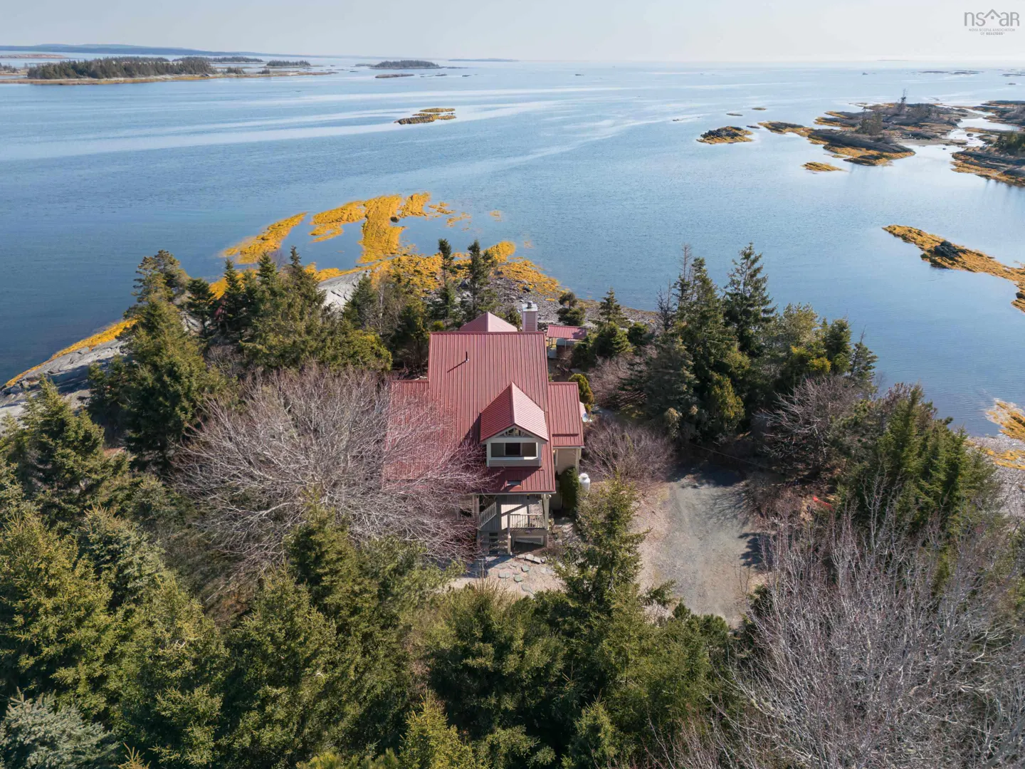 Aerial view of a house with a red roof, surrounded by trees, on a rocky island in a blue ocean.