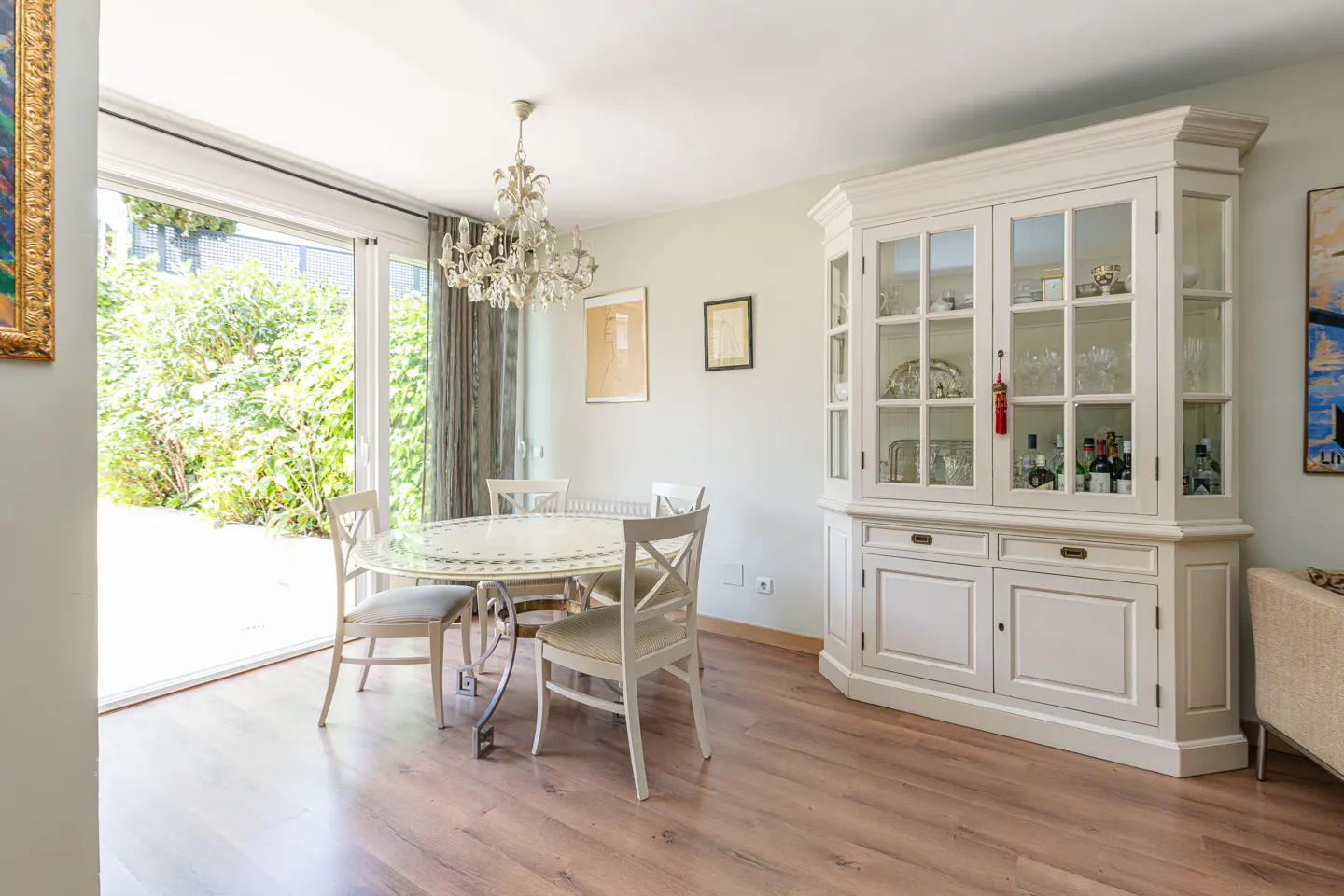Bright dining room with wood floors, a glass table, white chairs, and a white china cabinet filled with glassware and bottles. Sliding glass doors open to a green garden.