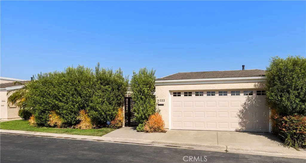 Beige single-story house with a two-car garage, green bushes, and a black metal gate under a clear blue sky.