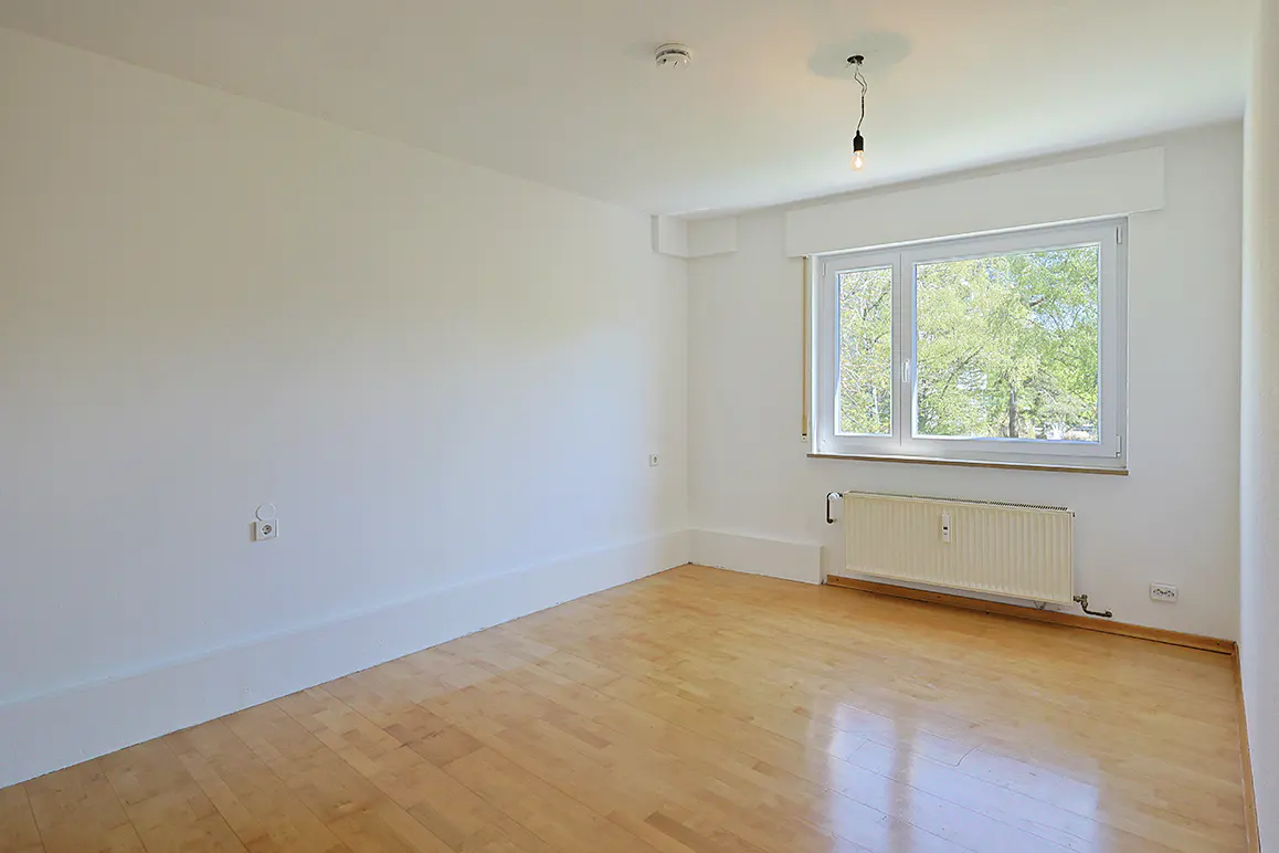 Empty room with white walls, light wood flooring, a window overlooking trees, and a white radiator below the window.