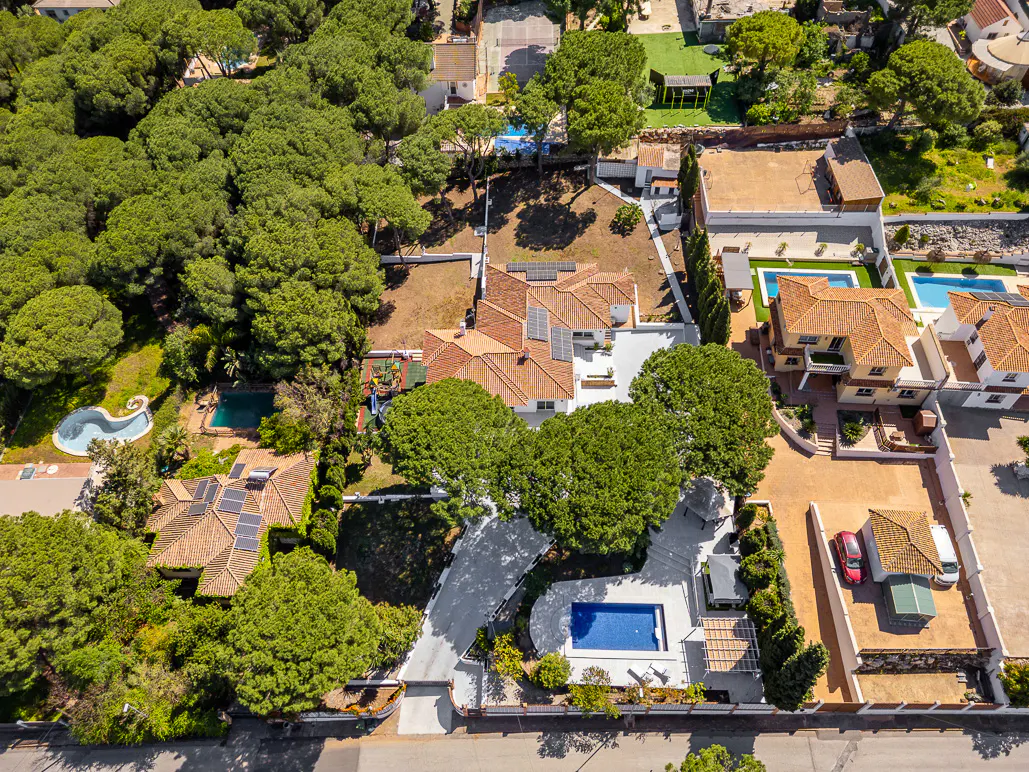Aerial view of luxury homes with pools, red tile roofs, and lush green trees. Solar panels are visible on some roofs.