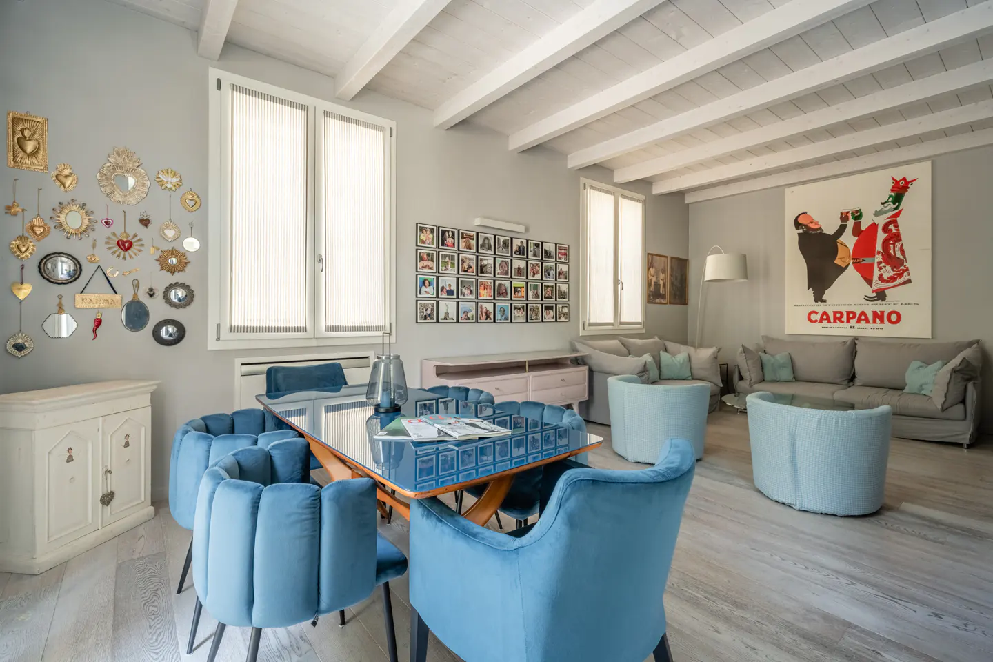 Bright, modern living room with blue velvet chairs around a glass table. White walls, wooden floors, and a beamed ceiling. Artwork and photos decorate the walls.